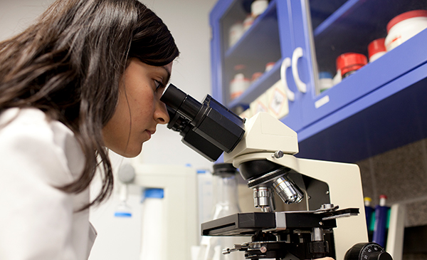 Female student wearing a white coat looking into a microscope