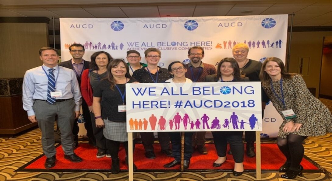 11 IDHD staff, faculty, and alumni pose in front of a sign which reads 