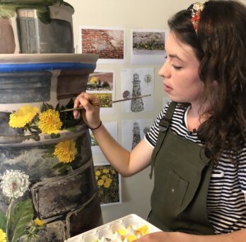 Kaitlin Stober paints yellow flowers on her lighthouse art installation piece
                  