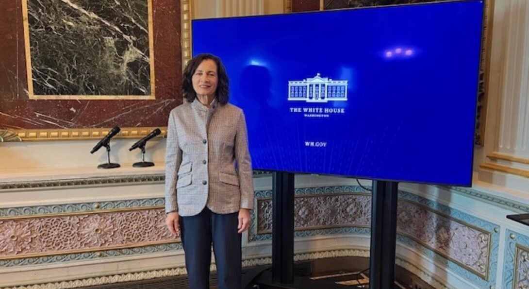 (ID: woman with dark hair wearing gray/black business suit standing next to large monitor showing white house logo.  Behind is shown marbled walls.